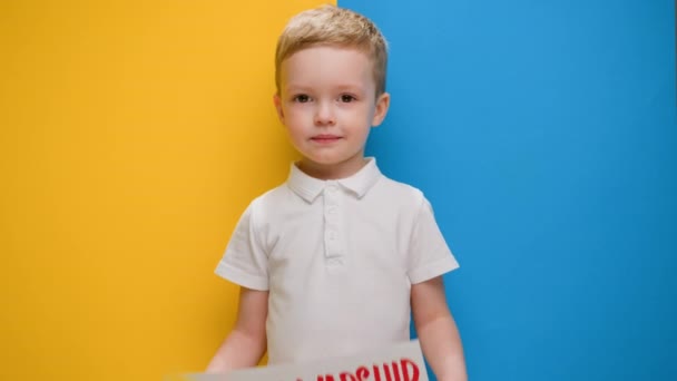 Portrait Blond little smiling boy, raises banner with inscription Russian warship go fuck your self standing on blue-yellow studio background. No war, stop war, russian aggression. — ストック動画