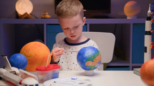 Blond boy paint planet solar system Earth with colorful paint sitting home table in evening, planet solar system, spaceships and space shuttle from constructor around. Cosmonautics Day on April 12. — Stock Photo, Image