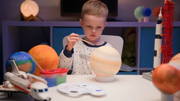 Blond boy paint planet solar system Saturn with colorful paint sitting home table in evening, planet solar system, spaceships and space shuttle from constructor around. Cosmonautics Day on April 12. — Stock Photo, Image