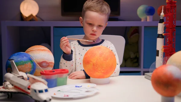 Menino loiro desenha estrela do sol com pintura multicolorida, sentado à mesa em casa à noite, planeta do sistema solar, naves espaciais e ônibus espacial do construtor ao redor. Dia da Cosmonáutica é 12 de abril. — Fotografia de Stock