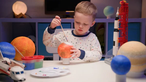 ブロンドの少年は、夜に家のテーブルに座ってカラフルな塗料で惑星の太陽系火星、惑星の太陽系、宇宙船や周りのコンストラクタからスペースシャトルをペイントします。4月12日の宇宙の日. — ストック写真