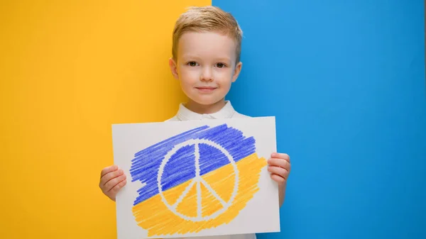 Pacifist child protests against war in Ukraine. Blond-haired boy smiling on yellow-blue background of flag of Ukraine holds banner with logo of anti-war movement Pacific. — Stock Photo, Image