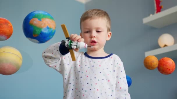 Blond-haired boy is playing with space satellite made by constructor, flying in space near planets solar system. Child dreams of becoming an astronaut to fly on shuttle space. Space day 12 April. — Stock Video