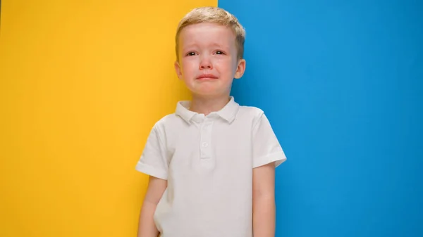 Blond boy is crying from heartache because of war in Ukraine standing on yellow-blue background of flag of Ukraine. Child against war, boy crying from war, Russian aggression in Ukraine. — Stock Photo, Image