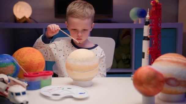 Loira menino pintar planeta sistema solar Saturno com tinta colorida sentado em casa mesa à noite, planeta sistema solar, naves espaciais e ônibus espacial do construtor ao redor. Dia da Cosmonáutica em 12 de abril. — Vídeo de Stock