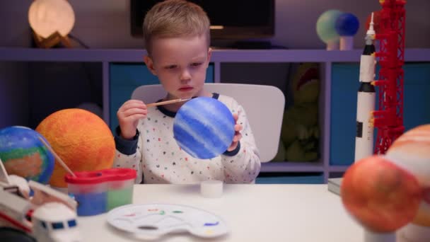 Blonďatý chlapec malovat planetu sluneční soustavy Neptun s barevnou barvou sedí doma stůl ve večerních hodinách, planeta sluneční soustavy, kosmické lodě a raketoplán z konstruktéra kolem. Den kosmonautiky 12. dubna. — Stock video
