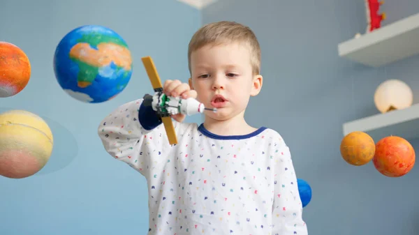 ポートレートブロンドの少年は、太陽系の近くの宇宙を飛んで、コンストラクタによって作られた宇宙衛星で遊んでいます。子供はシャトルスペースで飛行する宇宙飛行士になることを夢見ています。宇宙の日4月12日. — ストック写真
