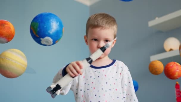 Menino loiro está brincando com foguete espacial de brinquedo, voando no espaço entre planetas do sistema solar. Criança está brincando em casa com foguete voando no espaço do sistema solar. Dia da Cosmonáutica em 12 de abril. — Vídeo de Stock