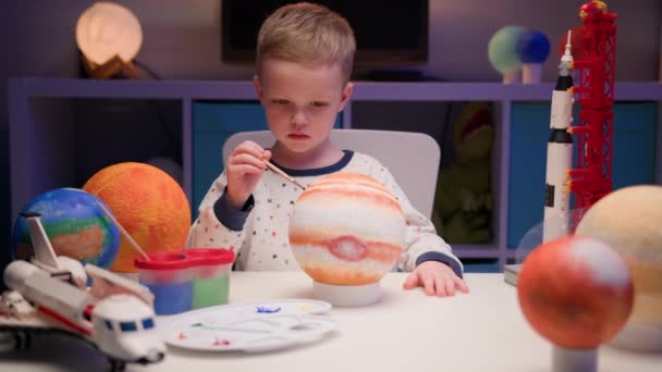 Blonder Junge bemalt Planet Sonnensystem Jupiter mit bunter Farbe am heimischen Tisch sitzend abends, Planet Sonnensystem, Raumschiffe und Raumfähre vom Konstrukteur herum. Tag der Kosmonautik am 12. April. — Stockvideo