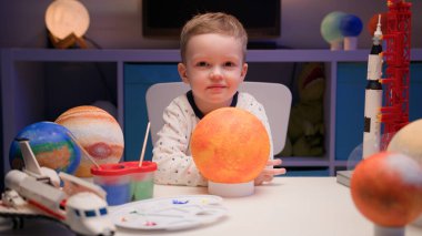 Portrait smiling little blond boy sitting at home at table near planet Sun around many planets of solar system. Child Studies Astronomy and Stars in Cosmonautics Day on April 12.