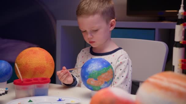 Blond garçon peindre planète système solaire Terre avec de la peinture colorée assis à la maison dans la soirée, planète système solaire, vaisseaux spatiaux et navette spatiale du constructeur autour. Journée Cosmonautique le 12 avril. — Video