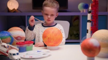 Sarışın çocuk güneş yıldızını çok renkli boyasıyla çiziyor, akşamları ev masasında oturuyor, güneş sistemi gezegeni, uzay gemileri ve uzay mekiği etrafta. Kozmonotik Gün 12 Nisan.