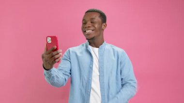 Cute smiling black man, African American 20 years old in denim shirt, answers video phone call, talks to his family on red smartphone, rushes his hand to bring interlocutor on pink studio background.