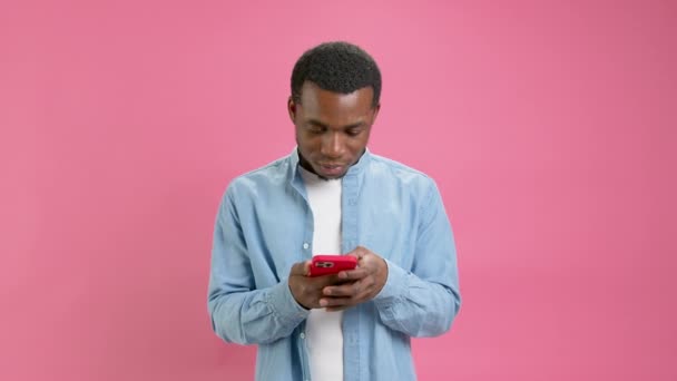 Shocked happy victory young African American 20 years old in denim shirt, emotion of winning casino or sports betting in mobile application poses in isolation on pink background. Emotions of victory. — стокове відео