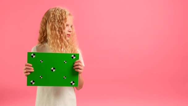 Ευτυχισμένο ξανθό κορίτσι κρατώντας Banner με πράσινο Screen Tracks σημεία για Copy Space. Κενό πράσινο πίνακα οθόνης. Χαμόγελα και κοιτάζει κάμερα με χώρο για κείμενο ή διαφήμιση σε ροζ φόντο. — Αρχείο Βίντεο