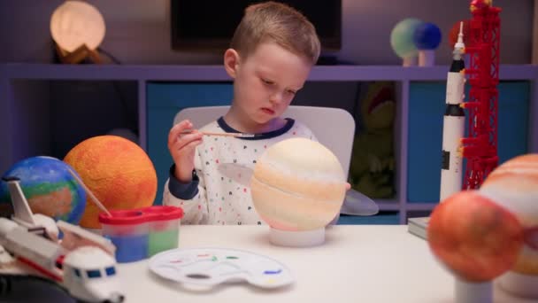 Niño rubio pintar planeta sistema solar Saturno con pintura de colores sentado mesa en casa por la noche, planeta sistema solar, naves espaciales y transbordador espacial de constructor alrededor. Día de la Cosmonáutica el 12 de abril. — Vídeo de stock