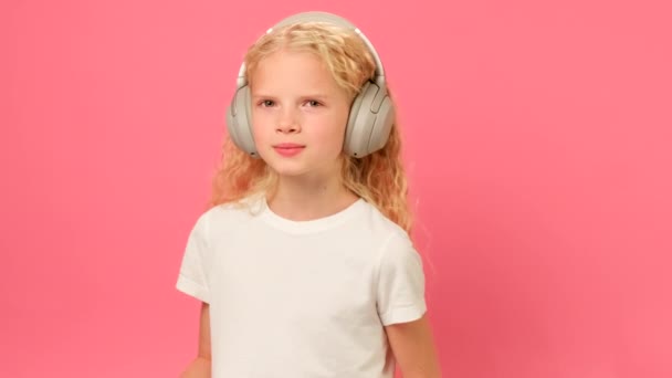 Portrait blonde teenage girl listening to her favorite music in gray headphones and looking at camera on pink studio background. Slow motion portrait girl enjoying listening to songs with headphones. — Αρχείο Βίντεο