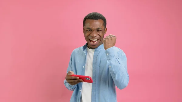 Chocado vitória feliz, jovem afro-americano 20 anos em camisa jeans é surpreendido agradável notícia olhando para o telefone, ganhando um aplicativo de casino ou apostar em esportes de pé no fundo do estúdio rosa. Imagens De Bancos De Imagens Sem Royalties