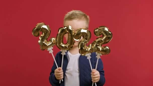 Cute blond Caucasian child boy holds golden inflatable numbers 2022 in his hands and covers his face with figures. Portrait of boy rejoicing incoming 2022 year tiger standing on red studio background. — Stock Video