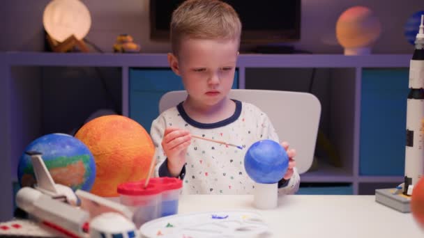 Niño rubio pintar planeta sistema solar Plutón con pintura colorida mesa de estar en casa por la noche, planeta sistema solar, naves espaciales y transbordador espacial de constructor alrededor. Día de la Cosmonáutica el 12 de abril. — Vídeo de stock