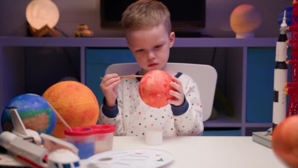 Blond garçon peindre planète système solaire Mercure avec de la peinture colorée assis à la maison dans la soirée, planète système solaire, vaisseaux spatiaux et navette spatiale du constructeur autour. Journée Cosmonautique le 12 avril. — Video
