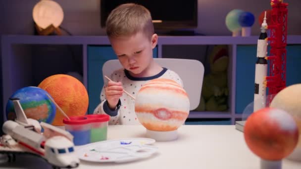 Biondo ragazzo dipingere pianeta sistema solare Giove con vernice colorata seduto a casa tavolo in serata, pianeta sistema solare, astronavi e navetta spaziale dal costruttore intorno. Giorno di Cosmonautica il 12 aprile. — Video Stock