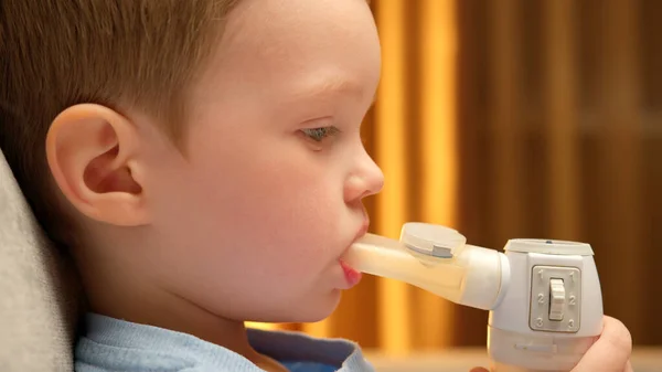 Blond Caucasian child with asthma problems inhales with tube in his mouth. Child has respiratory infection or bronchitis, and he is breathing heavily. Treatment of pneumonia with nebulizer at home.