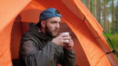 Slow motion portrait bearded traveler man sitting in an orange tent wearing blue cap and gray jacket in nature and drinking cup of hot tea or fragrant coffee, looking into camera and smiling.