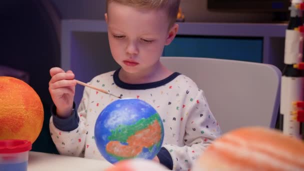 Blonďatý chlapec malovat planetu sluneční soustavy Země s barevnou barvou sedí doma stůl ve večerních hodinách, planeta sluneční soustavy, kosmické lodě a raketoplán z konstruktéra kolem. Den kosmonautiky 12. dubna. — Stock video