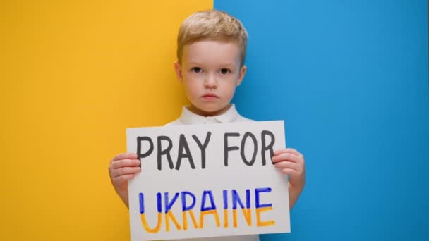 Portret Blond kleine jongen, verhoogt spandoek met opschrift Pray For Ukraine staan op blauw-gele studio achtergrond. Geen oorlog, geen oorlog, Russische agressie.. — Stockvideo