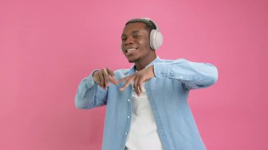 Portrait rapper cheerful African-American man in denim shirt and Bluetooth headphones on his head listens to music, smiles with pleasure and raps to hip-hop rhythms on pink background.
