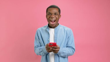 Şok olmuş mutlu zafer, 20 yıllık kot gömlekli genç Afrikalı Amerikalı telefona bakmak, kumarhane başvurusu kazanmak ya da pembe stüdyo arka planında spor bahisleri oynamak hoş bir haber..