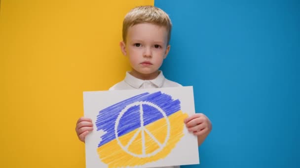 Ξανθός-μαλλιά αγόρι σε κίτρινο-μπλε φόντο της σημαίας της Ουκρανίας κατέχει πανό με το λογότυπο του αντιπολεμικού κινήματος του Ειρηνικού. Ειρηνιστικό παιδί διαμαρτύρεται κατά του πολέμου στην Ουκρανία. — Αρχείο Βίντεο