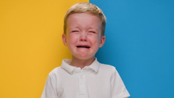 Retrato pequeño niño rubio llorando sobre la bandera de fondo amarillo-azul de Ucrania. Crisis en Ucrania, guerra contra los niños, agresión rusa catástrofe humanitaria, asistencia a los niños en Ucrania. — Vídeo de stock