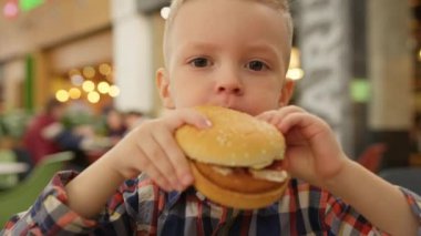 Blond caucasian 4-year-old boy eats hamburger with beef in fast food restaurant. Child bites cheeseburger on camera and eats. Child eats junk food in fast food.