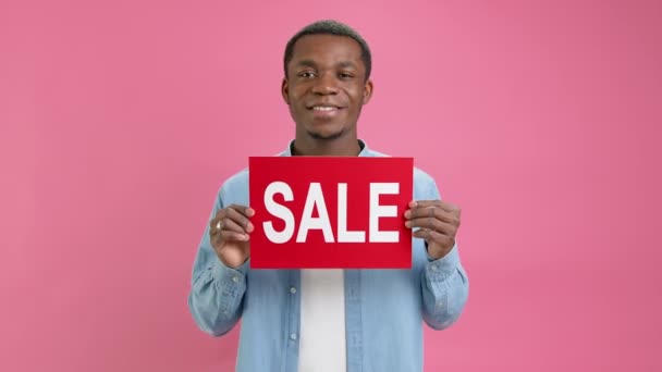 Happy African American man in denim shirt raises banner with inscription sale, waves his head approvingly, approves a discount and sale at low prices with place for layout on pink studio background. — Video
