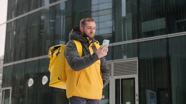 Correio sem barba com mochila amarela em suas costas em uniforme caminha rua abaixo no inverno entre edifícios de vidro altos, navega cidade por telefone, e encontra endereço de entrega. Conceito de entrega de correio. Fotos De Bancos De Imagens