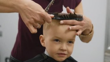 Process of hairdresser cutting hair. Blond caucasian boy is sitting in barber shop and hairdresser girl is cutting his hair. Child gets fashionable haircut, hairdresser cuts child hair with scissors.