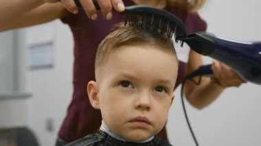 Blow-drying hair for little boy, hair styling, fashionable haircut in barber shop by master stylist. Close-up of process of drying hair of child in barber shop.