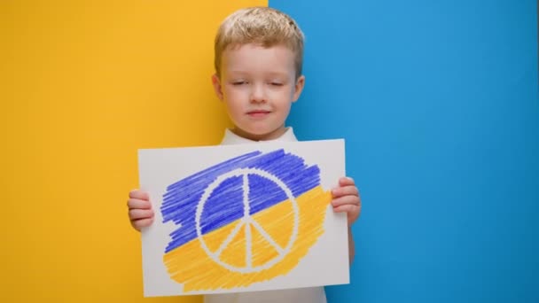 Niño rubio sonriendo sobre fondo amarillo-azul de la bandera de Ucrania sostiene pancarta con el logotipo del movimiento anti-guerra Pacífico. Protestas infantiles pacifistas contra la guerra en Ucrania. — Vídeos de Stock