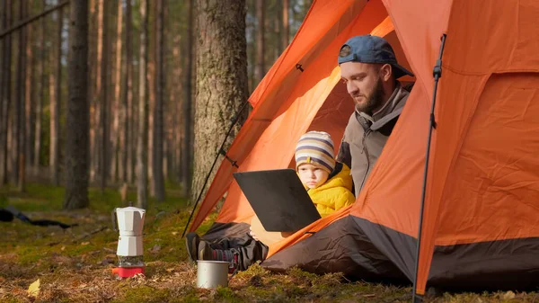 Father and son relaxing together in nature in the spring forest, sitting in an orange tent and watching videos together on laptop screen, communicating via video chat link via notebook with relatives.