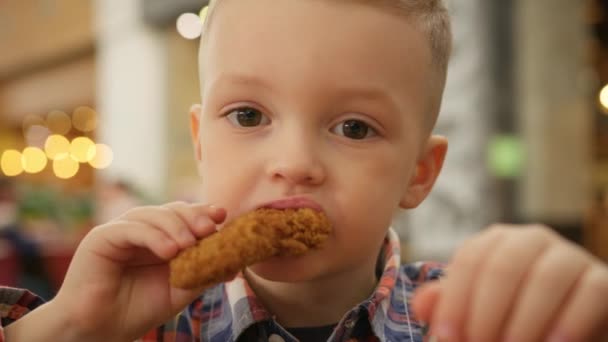 Blond Caucasian boy eats deep-fried chicken close-up. Portrait child eating chicken nuggets, chicken leg, chicken fillet from a fast food restaurant in a fast food restaurant. — ストック動画