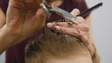 Blond caucasian boy is sitting in barber shop and hairdresser girl is cutting his hair. Child gets fashionable haircut, hairdresser cuts child hair with scissors. Close up of hairdresser cutting hair.