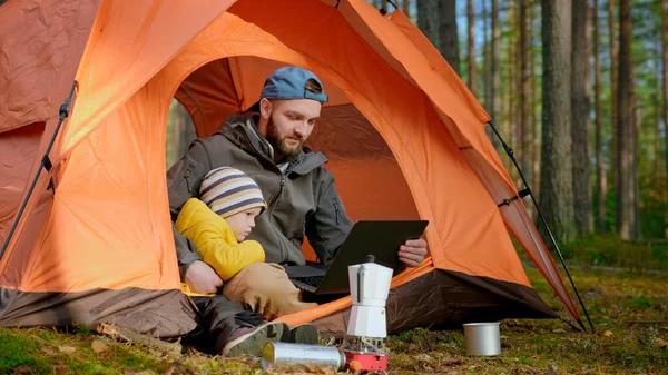 Father and son relaxing together in nature in the spring forest, sitting in an orange tent and watching videos together on laptop screen, communicating via video chat link via notebook with relatives.