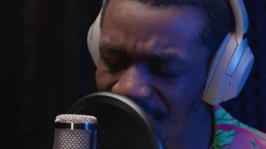 Portrait black person singer in headphones recording a music track in professional recording studio. Man sings rap into microphone with headphones on his head emotionally waving his hands.
