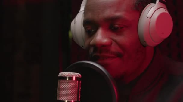 Black man singer in headphones recording music track in professional recording studio. Man sings rap song into microphone with headphones on his head against background noise-absorbing foam rubber. — Video
