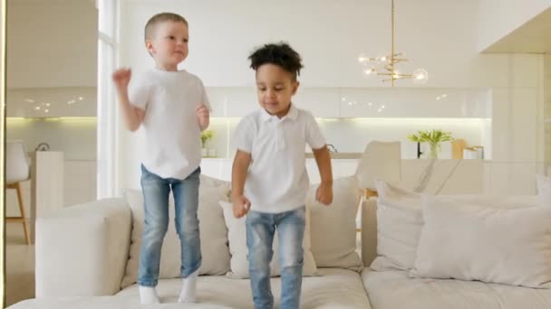Caucasian blonde child and African boy have fun playing jumping on couch in room at home. Two children in white polos and jeans play together, jump on couch, happy friendship. — Vídeo de Stock