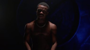 Cool African American teenager with bare, pumped-up body sings hip-hop song and emotionally gestures with his hands. Man raps in music video and sings rap song against blue underground background. 