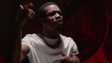 Black man teenage hip hop artist raps and waves his arms while recording music video for song. Portrait black African American man hip hop singer singing rap song in white t-shirt on red background.