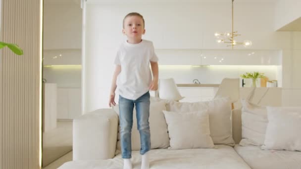 Happy blonde caucasian child has fun jumping up and down on light sofa. Child enjoys carefree childhood by jumping on bed like on trampoline at home. Jump on couch. — Stok video